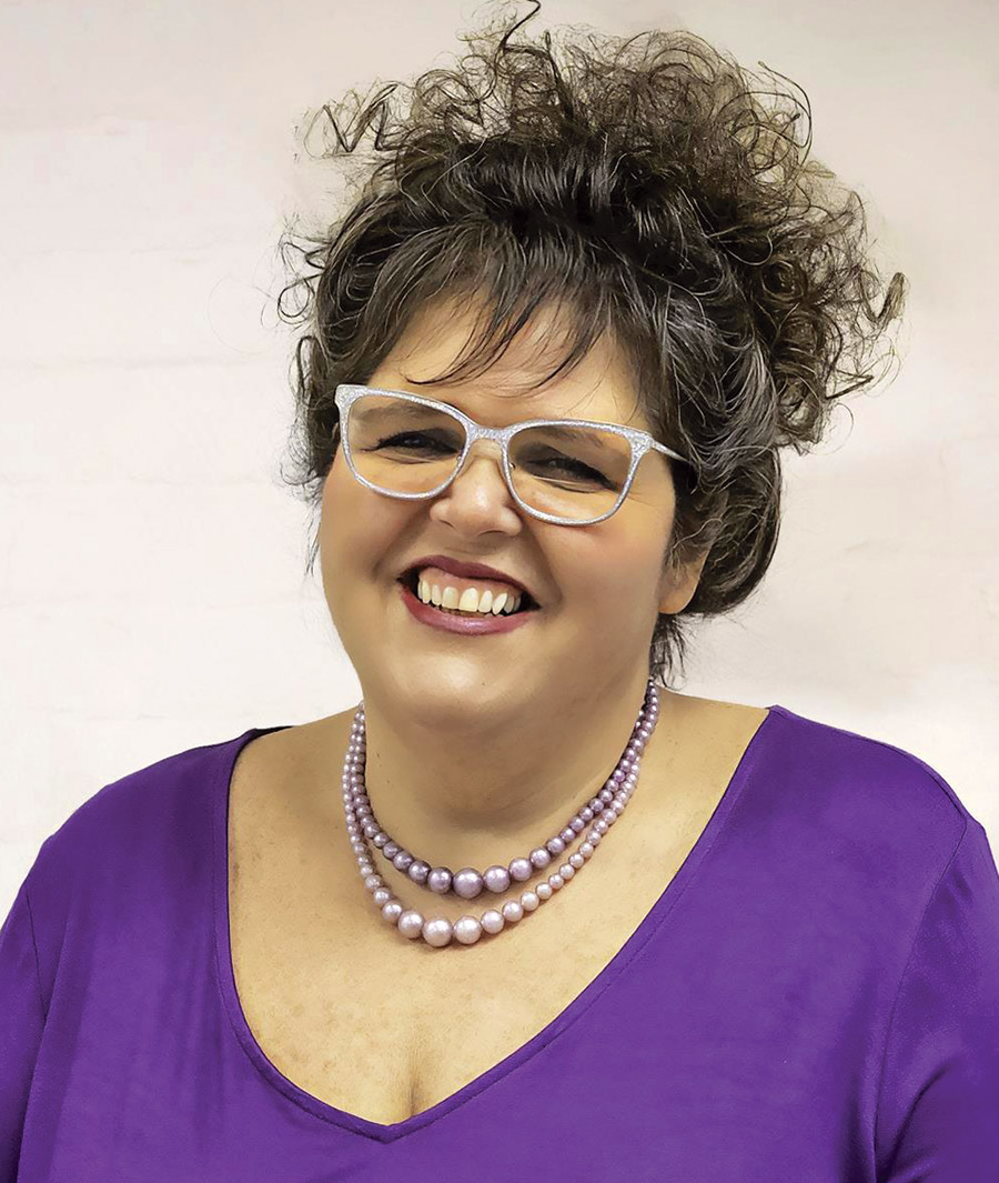Michelle Bannon smiles and poses for a picture in her chrome colored prescription see-through glasses and purple blouse with purple colored pearl necklaces