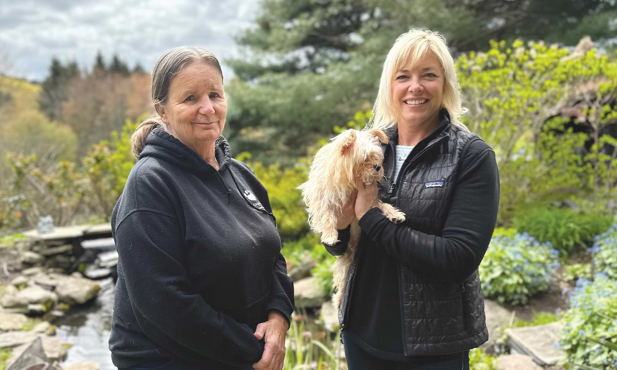 Marge Bart MBA ’85 smiles beside Cindy Charnetski ’97 who holds the rescue dog Mario, with the verdurous green of the Blue Chip Farm Animal Refuge in the background