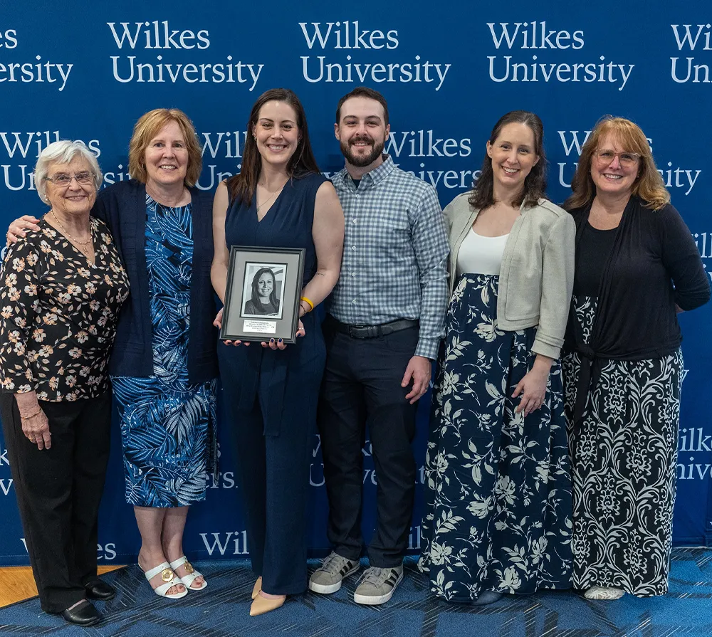 Six people smiling while holding a framed photo of a woman, standing in front of a blue banner that says 'Wilkes University'.