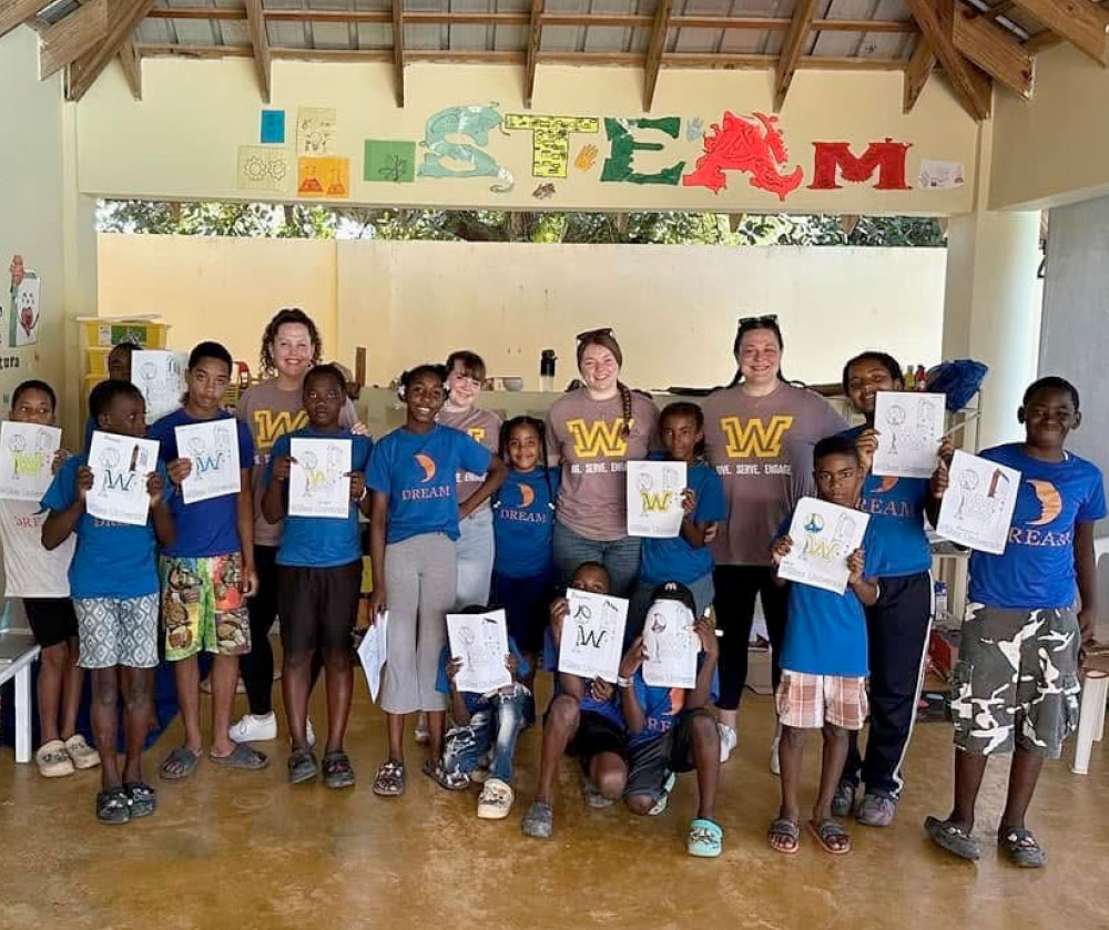 A group of children and adults pose for a photo while wearing brown shirts with a yellow 'W' and the text "WE SERVE. WE ENGAGE," and others wearing blue "DREAM" shirts, stand behind the children. The children, mostly wearing blue shirts, are holding up drawings they have created. On the back wall, there are colorful decorations spelling out "STEAM".
