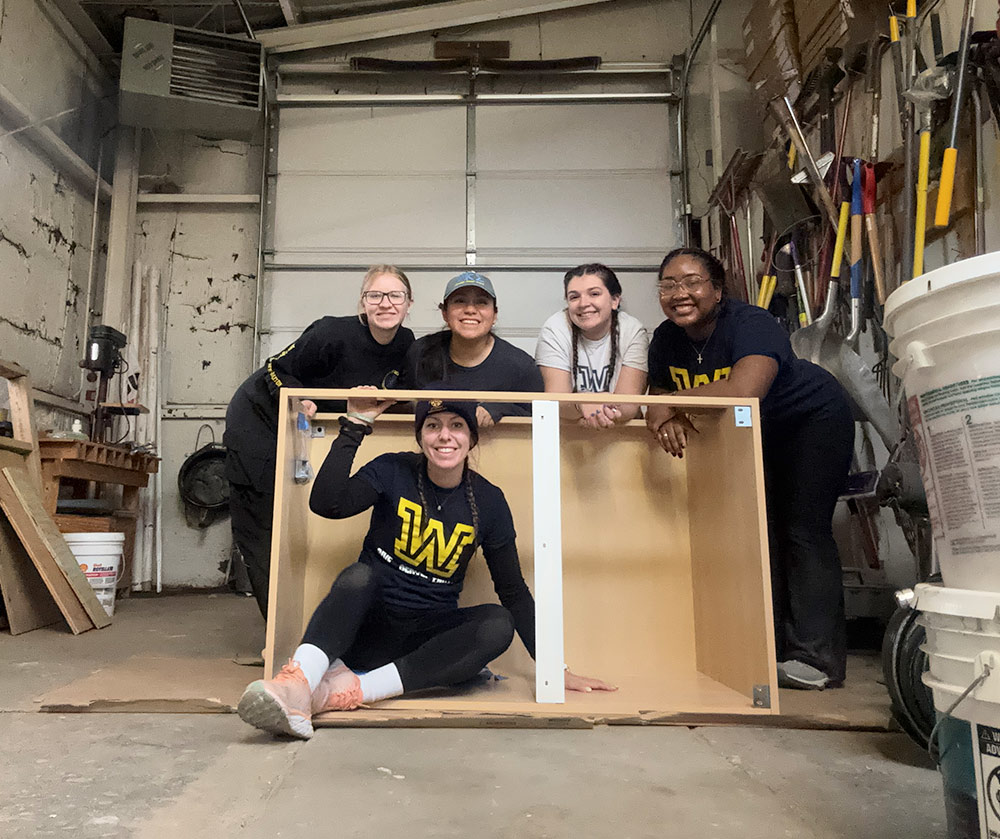 Five Wilkes University students smile for a photo while assembling a wooden shelf or cabinet in a workshop. 