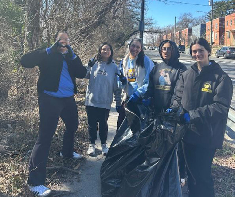 A group of five students smiles while holding a large black trash bag on the side of a road. They are participating in a cleanup and wearing gloves.