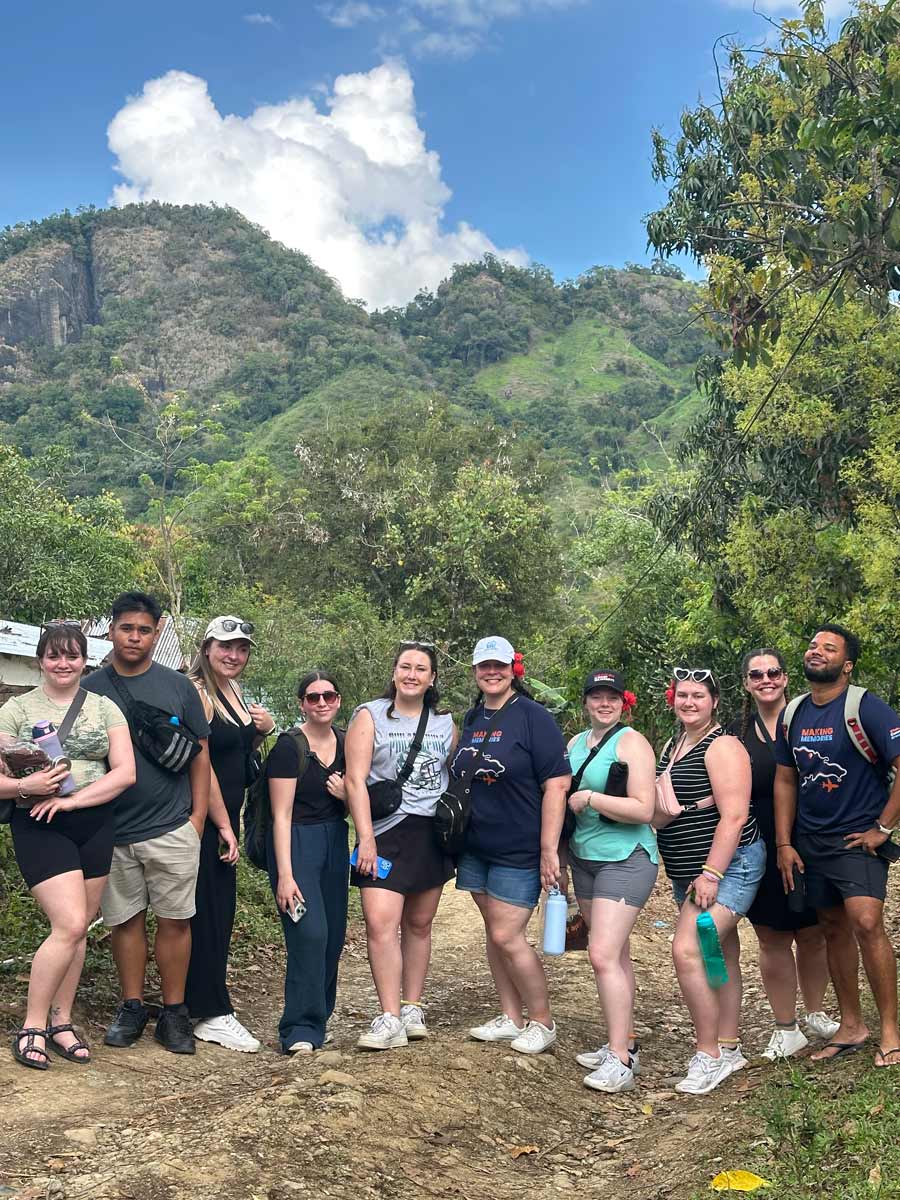 A group of ten smiling students stands on a dirt path in a lush, mountainous outdoor area. The students are casually dressed and are posing for a photo. A steep, rocky hill is visible in the background.