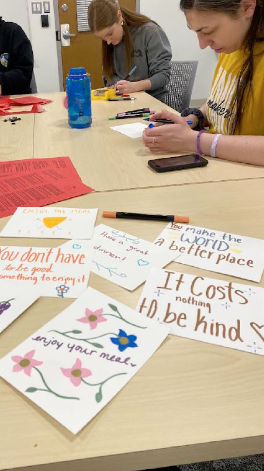 Students write and draw on small cards with messages like "Enjoy your meal" and "It costs nothing to be kind." A blue water bottle and markers are on the table.