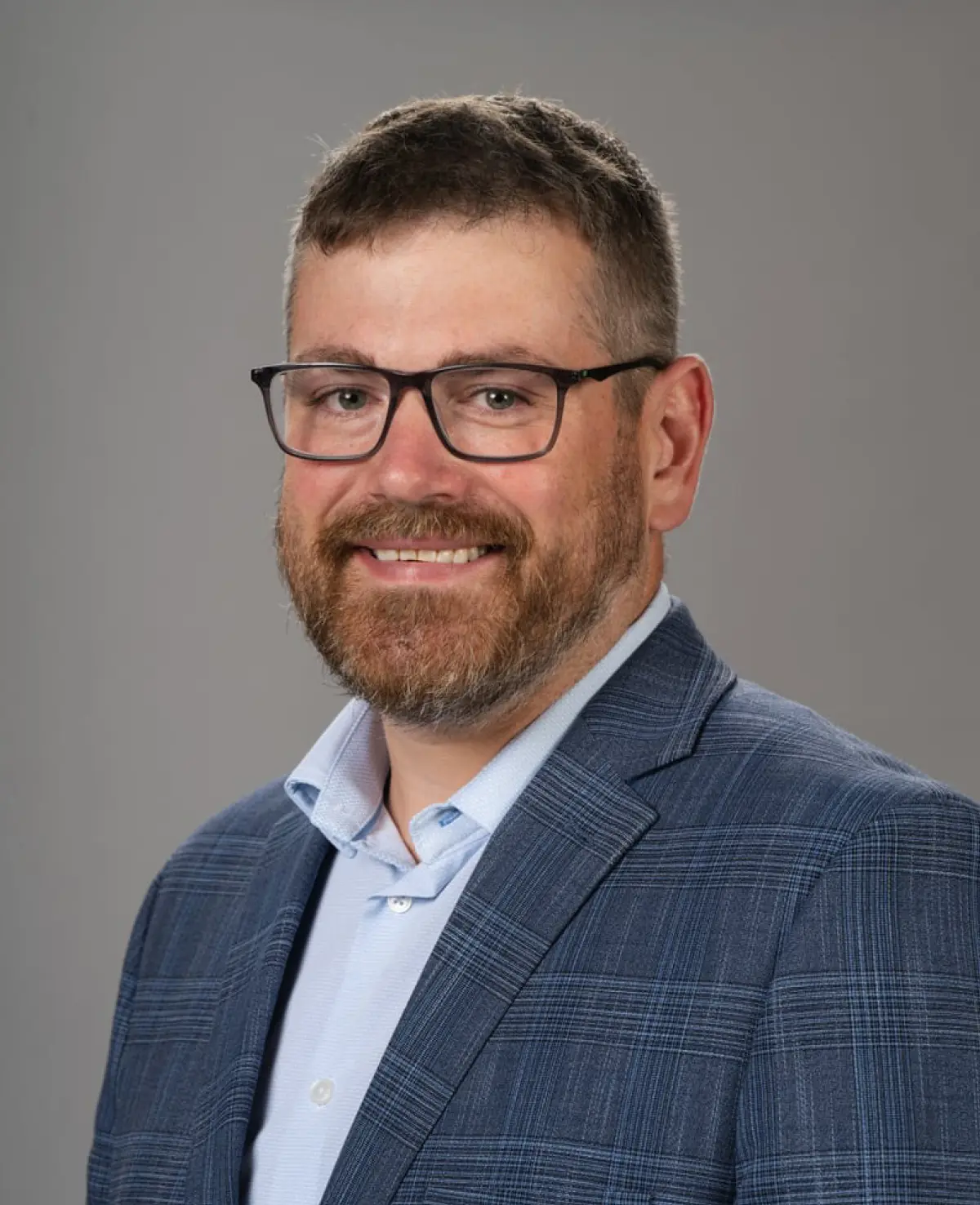 Portrait orientation photograph close-up headshot of Matt Crouse, a smiling man with black outer-frame prescription eyeglasses, a dark brown/grey beard, and dark brown hair as he is wearing a plaid dark marine blue business suit blazer with a light sky blue button-up dress shirt underneath