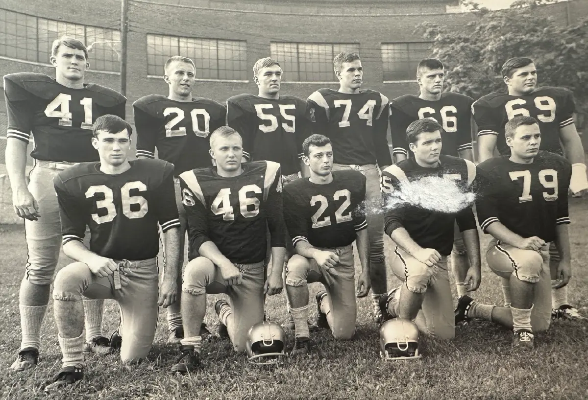 Landscape orientation black and white vintage photograph of Wilkes College's football team; Eleven young men in total in dark jerseys with numbers are posing in two rows in a kneeling position on a grassy field in front of a brick building outdoors; Two helmets belonging to two players are situated sitting on the grass directly in front of them; The photo has a visible tear or scuff mark over the chest of one of the kneeling players; William Stinger or affectionately known as Bill Stinger happens to be the football player pictured as #20