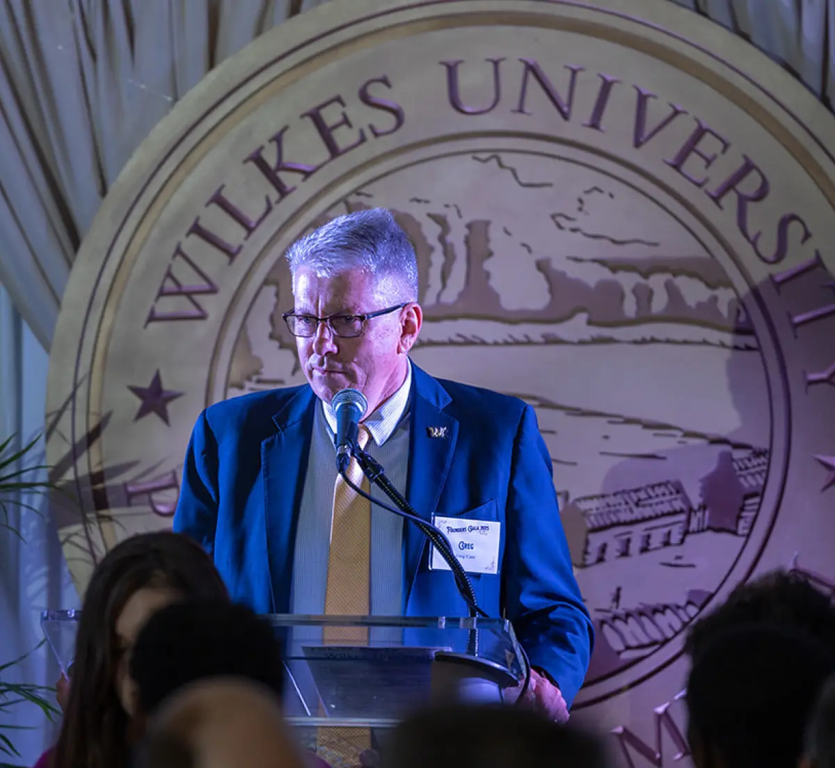 Cropped portrait orientation close-up photograph view of a man with gray hair and prescription eyeglasses wearing a dark blue colored business suit blazer jacket, white button-up dress shirt underneath, and a gold colored tie is speaking at a podium; This person happens to be President Greg Cant of Wilkes University, in which he is sharing remarks at this year’s Founders Gala; He is standing in front of a large, customized stylized circular seal emblem that says WILKES UNIVERSITY; In the background are beige colored curtains and in the foreground shows the backs of several people's heads who are seated at tables