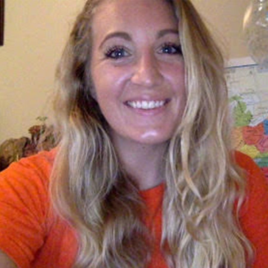 Headshot of Danielle Bartholomew, a woman with long, wavy blonde hair smiling at the camera. She is wearing a bright orange shirt, and a map is partially visible on the wall behind her.