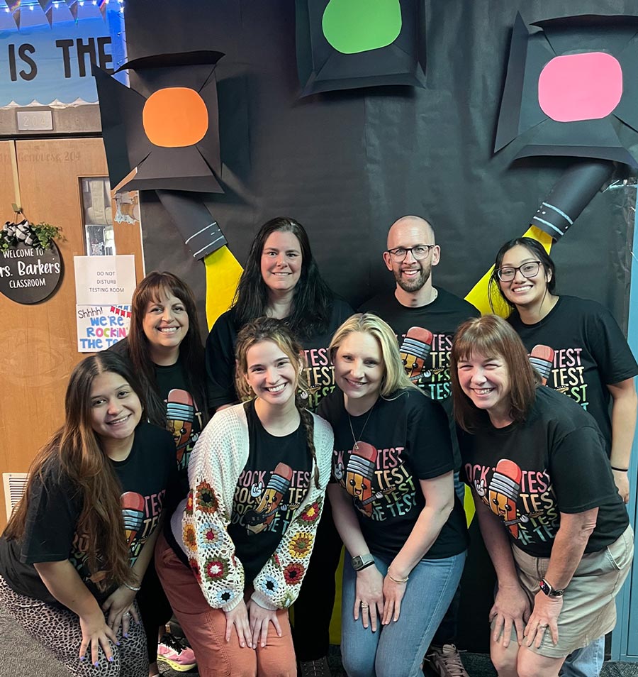 A group of eight educators, including Rosalinda Sosa in the back row on the right, pose together in a decorated classroom. They are all wearing matching black t-shirts with a pencil design that reads "Rock the Test" or "Test." Classroom decorations, including large black pencils with orange and green erasers, are visible in the background.