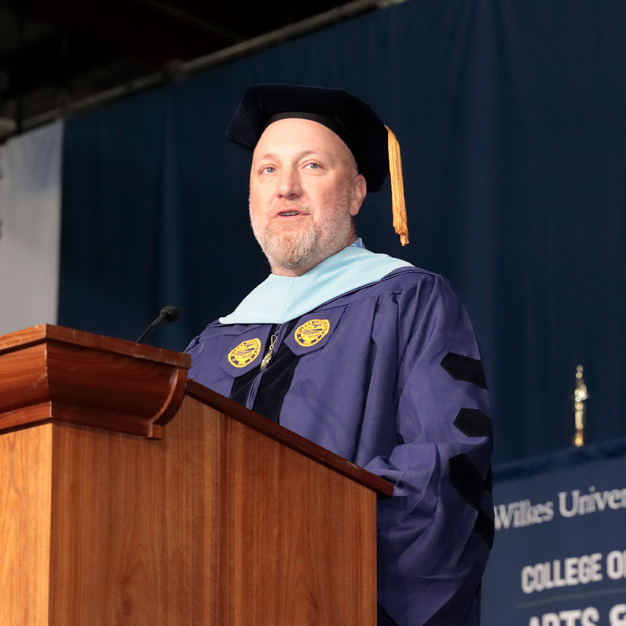 Dan Magie, a man with a beard, stands at a wooden podium and speaks. He wears a purple and light blue academic gown with a black mortarboard. In the background, a dark blue curtain is visible.