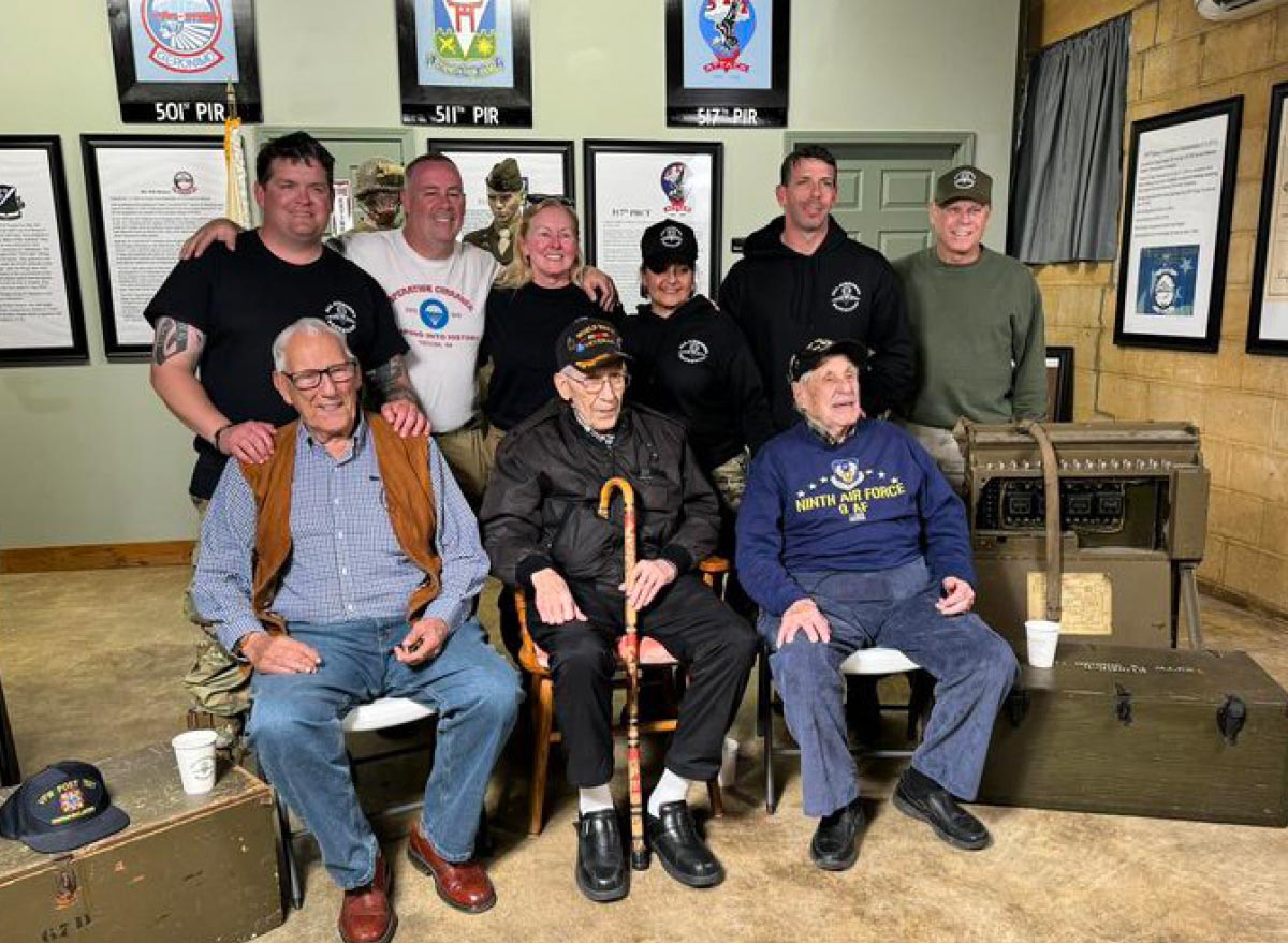 Sean Lockhead and members of the All Airborne Battalion pose with three WWII veterans in a room featuring 501st, 511th, and 517th PIR unit insignias.