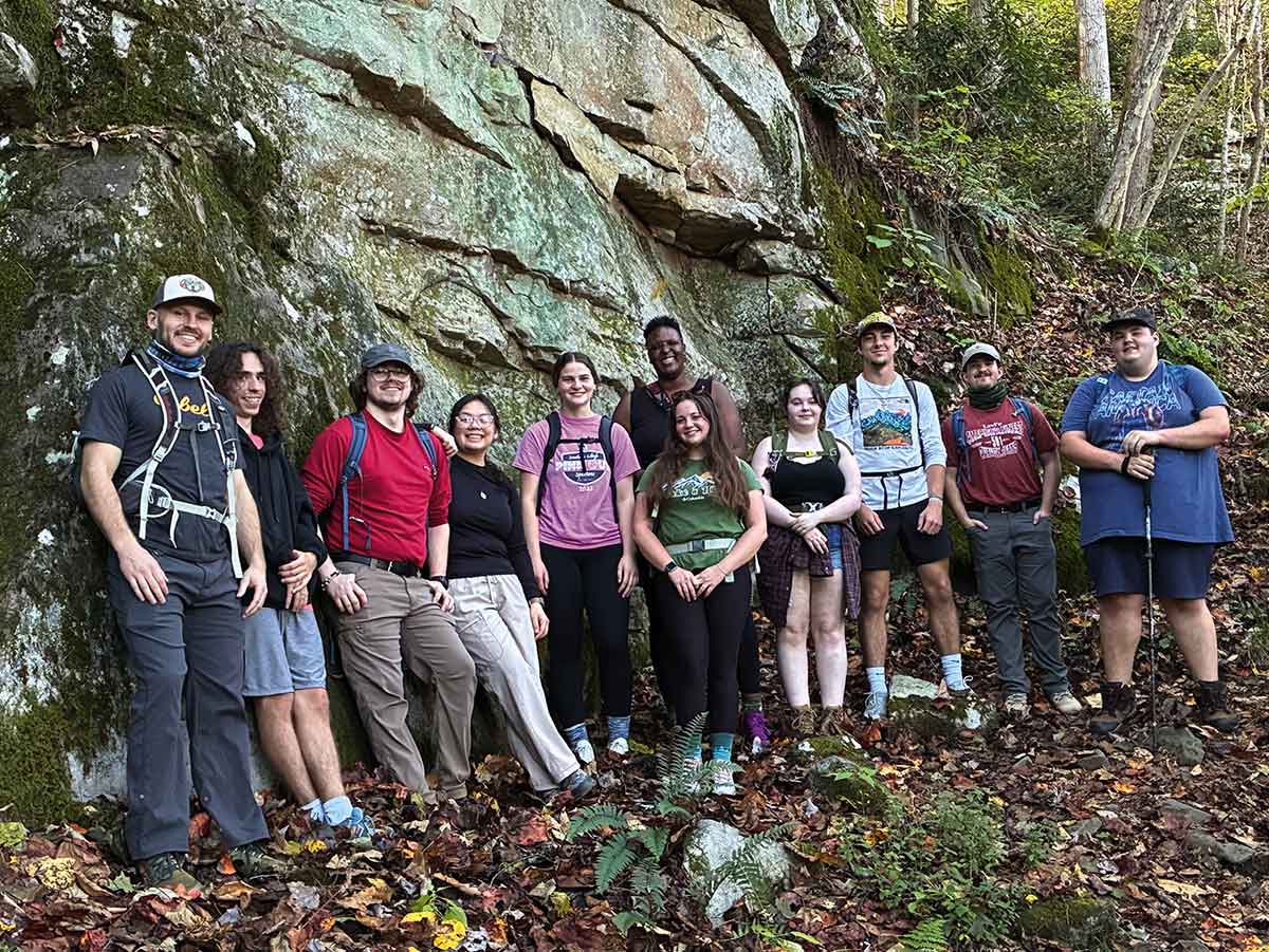 A group of ten young hikers poses for a photo against a large, moss-covered rock face on a forest trail.