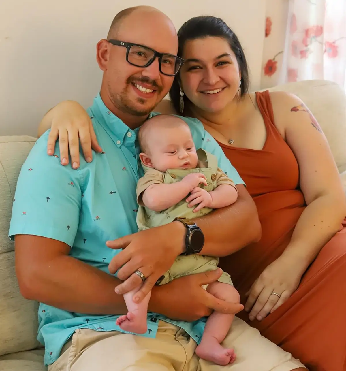 Christina (Harrison) Luksic and her husband smiling holding their first newborn baby as they are seated on a living room couch