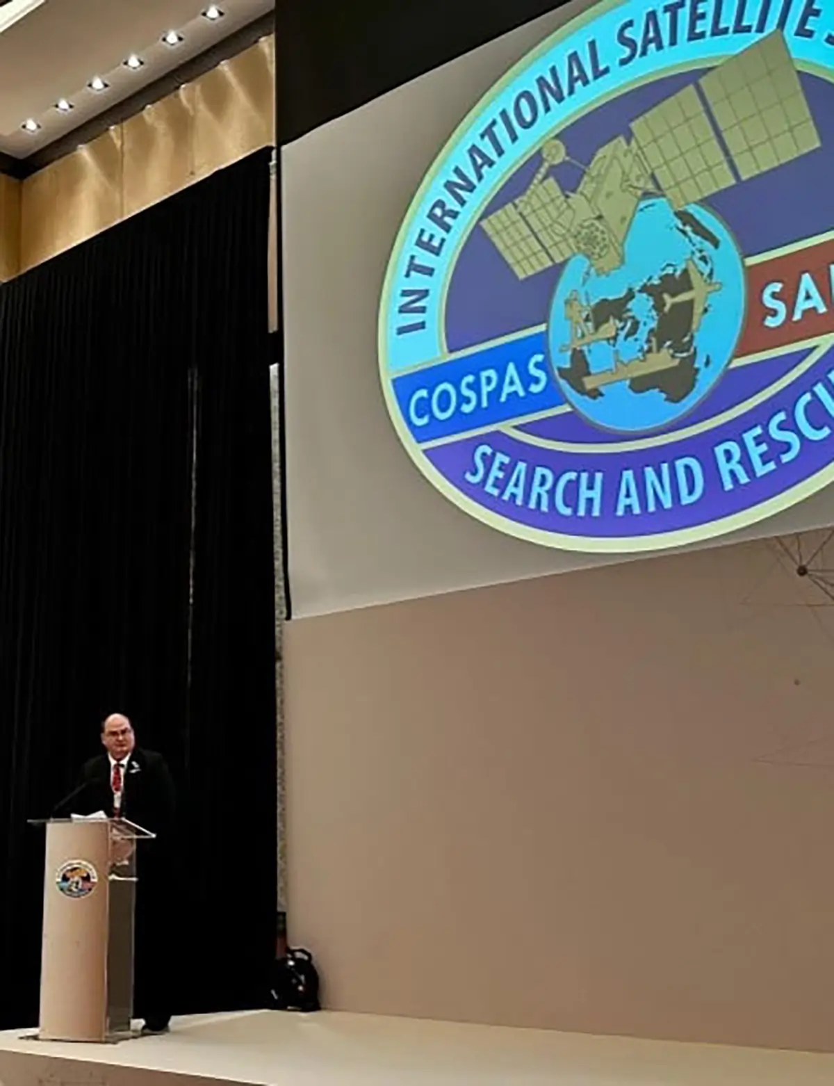 Allan Knox speaking at a podium standing in front of a projector screen that reads International Satellite COSPAS-SARSAT Search and Rescue