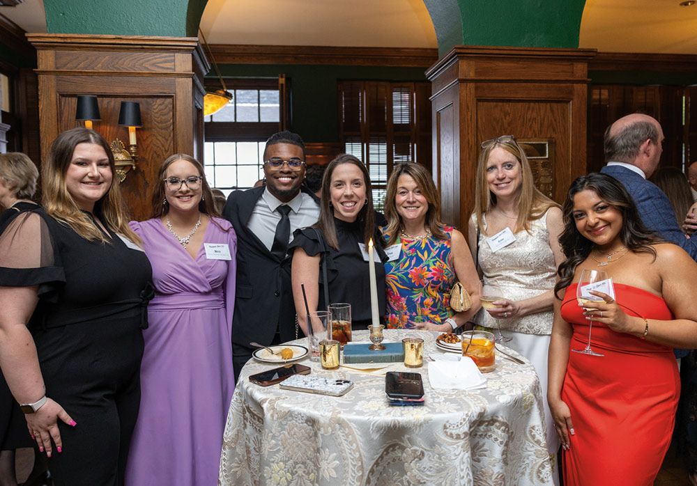 A diverse group of seven elegantly dressed students and guests pose together around a high-top table during a formal Wilkes University reception in a wood-paneled room.