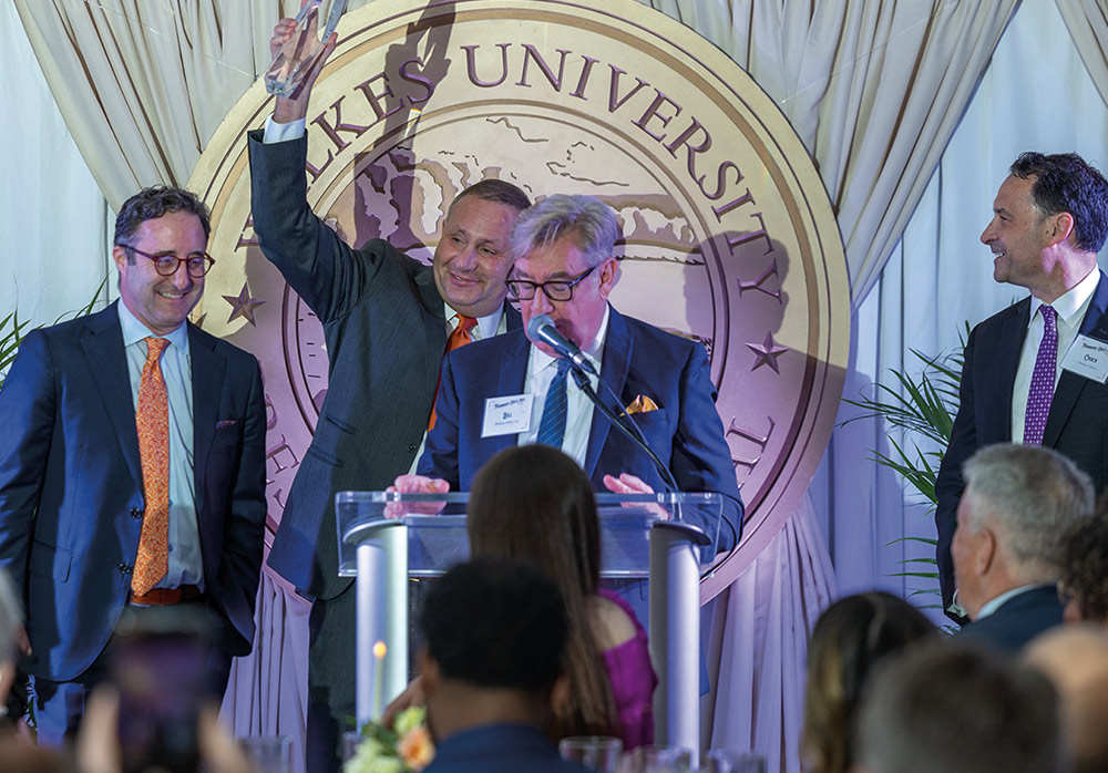 A man at a podium holds a glass award high in the air while three other men in suits smile and applaud in front of a large Wilkes University seal during a formal ceremony.