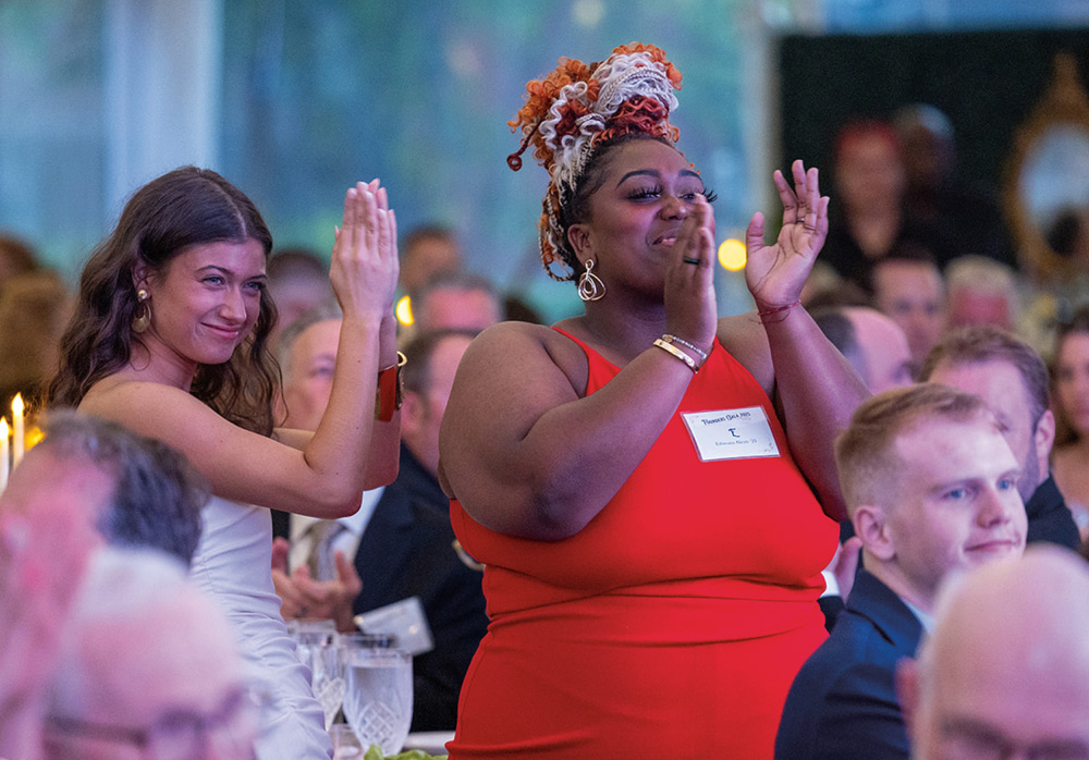 Guests at a formal dinner, including a woman in a vibrant red dress with stylized hair, smile and clap enthusiastically while seated at their tables during a gala event.