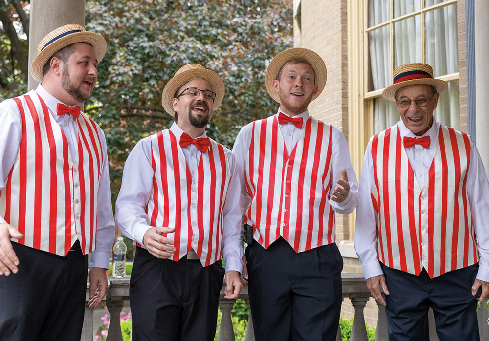 A barbershop quartet wearing matching straw boater hats, red bowties, and red-and-white striped vests performs outdoors on a stone balcony for homecoming guests.