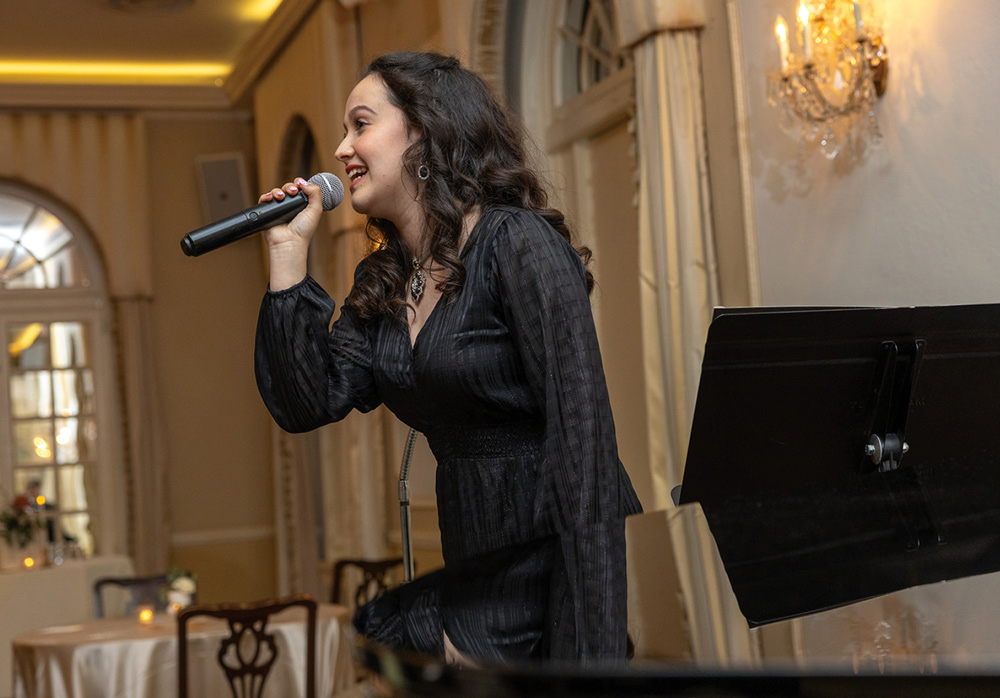 A student with long dark hair performs a song into a microphone at a formal event, standing near a piano in a ballroom with ornate lighting and neutral-toned decor.