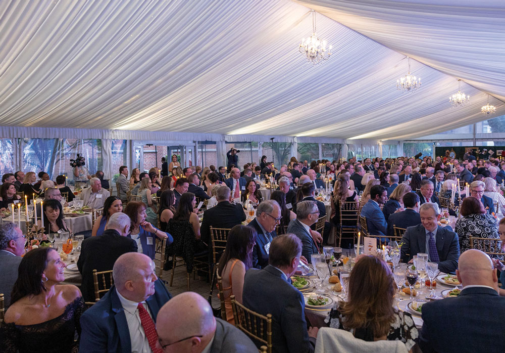 A wide-angle view of a large, crowded formal dinner taking place under a massive white event tent decorated with elegant chandeliers and candlelit table settings.