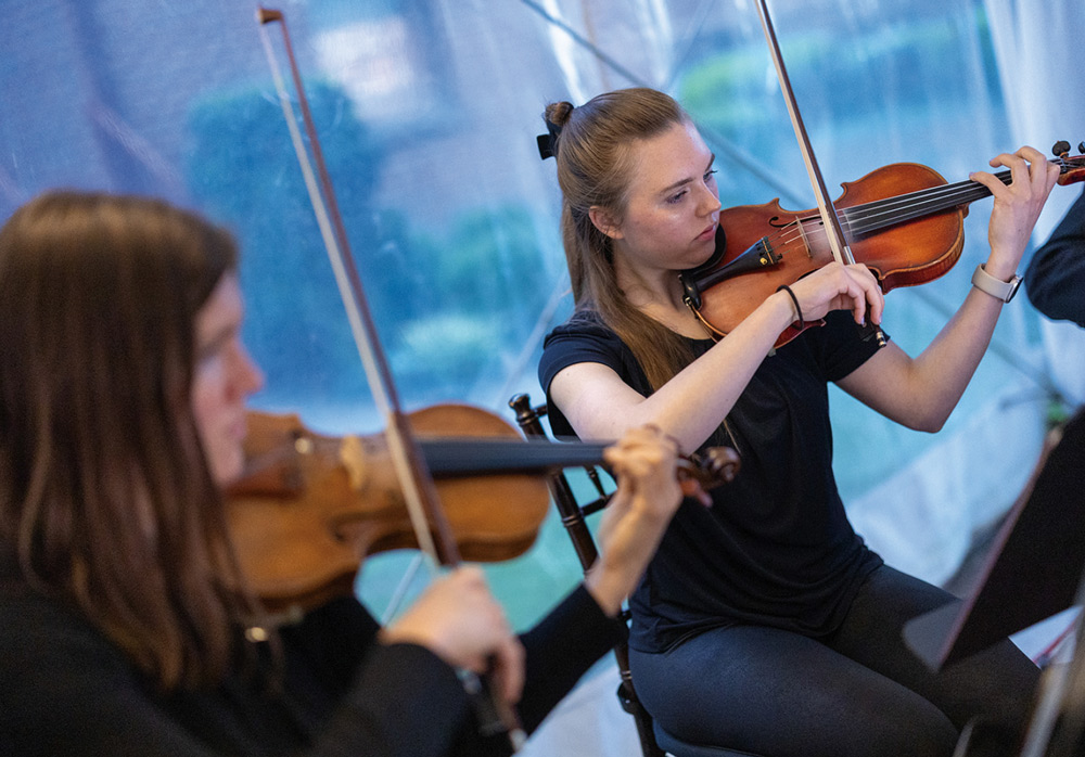 A close-up, candid shot of two student musicians focused on playing their violins during a formal event, with the outdoor evening light visible through a tent wall.