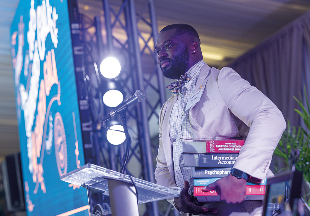 A man in a light-colored suit and bowtie stands at a podium carrying a large stack of textbooks while speaking at a formal Wilkes University gala event.