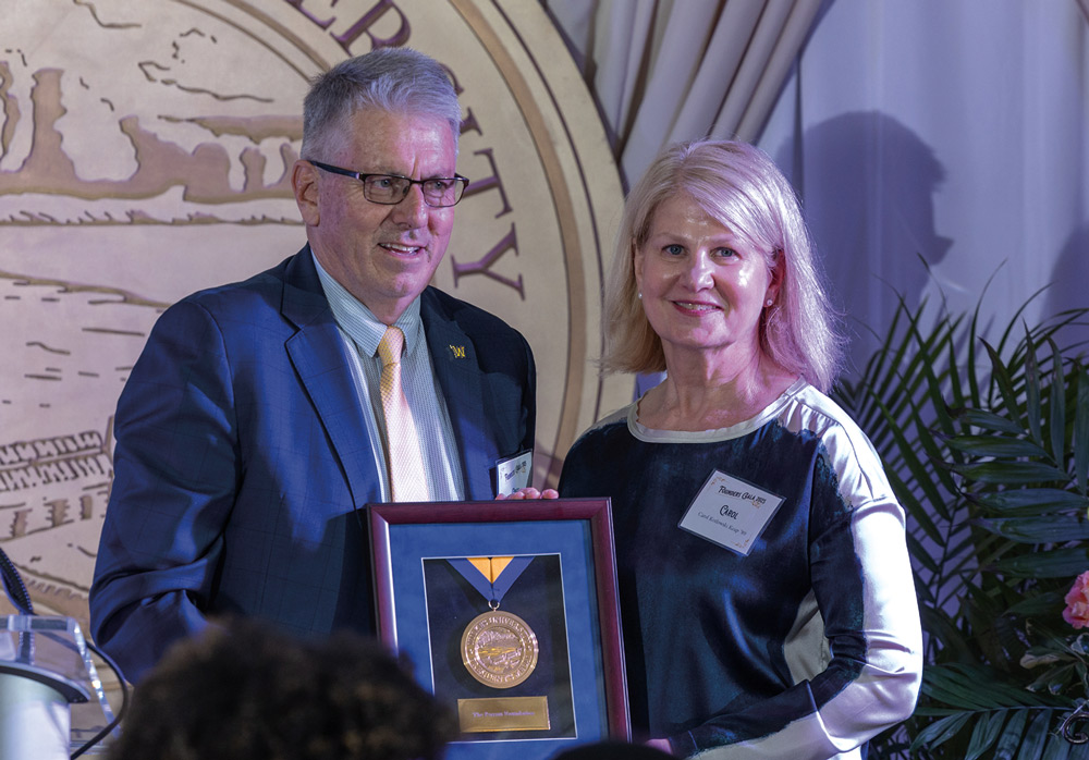 A man and woman pose together on stage holding a framed gold medal award in front of a large Wilkes University seal background during an official ceremony.