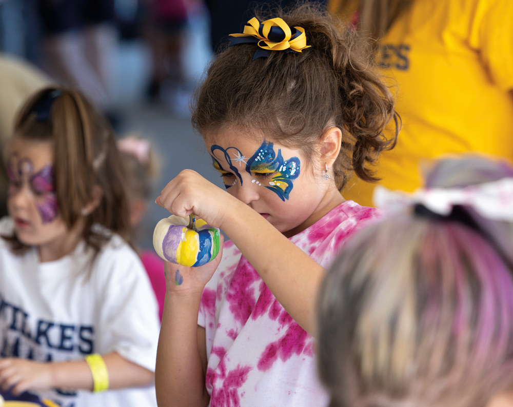 A young girl with a blue butterfly painted on her face focuses intently on painting a small pumpkin at a Wilkes homecoming event, while other children participate nearby.