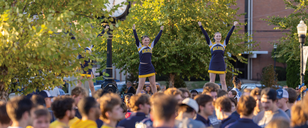 Wilkes University cheerleaders in navy and gold uniforms perform stunts with their arms raised for a large crowd of students gathered on campus during a sunny afternoon.