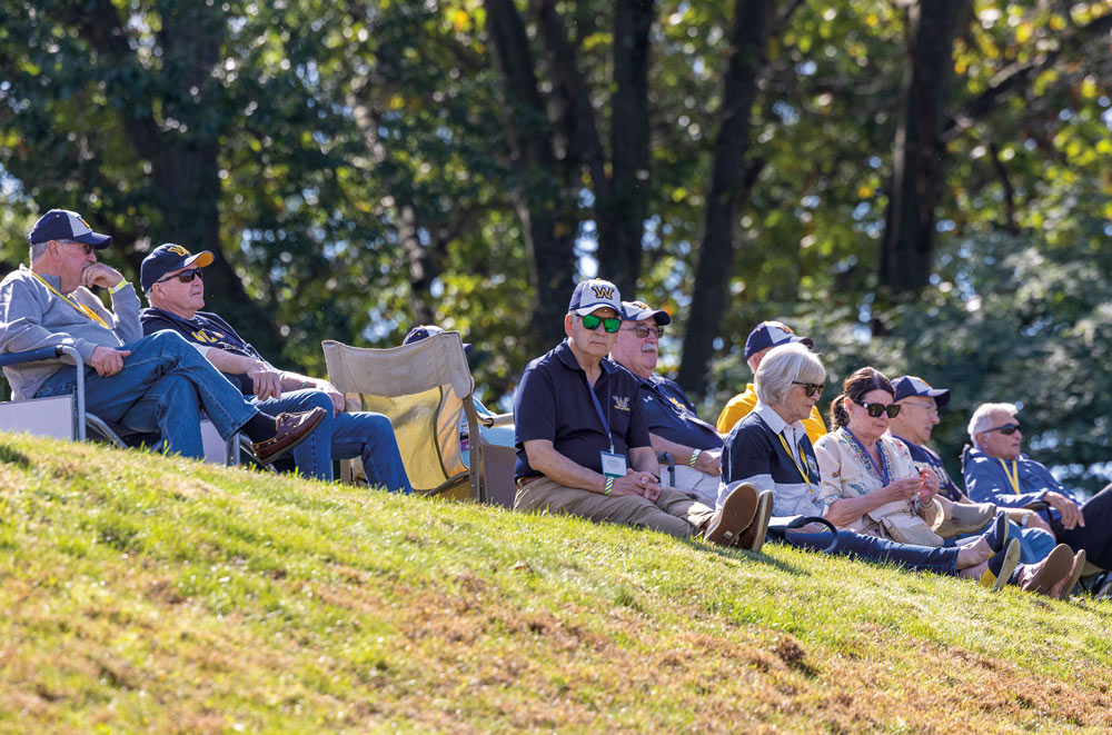 A group of Wilkes University alumni wearing matching "W" baseball caps sit in lawn chairs on a grassy hill, enjoying a clear, sunny day during the homecoming festivities.