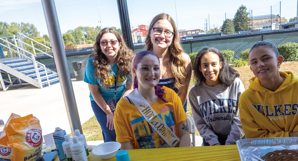 Five smiling Wilkes University students pose together behind a table. One student in the center wears a "Homecoming Court" sash, and event supplies are visible in the foreground.