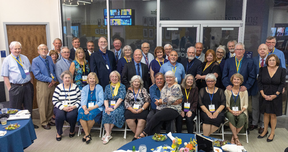 A large group of Wilkes University alumni and faculty pose for a formal group photo in an art gallery setting, many wearing name tags and "Wilkes" lanyards around their necks.