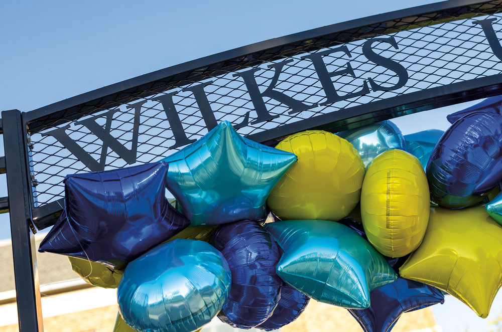 A close-up shot of the "Wilkes University" metal gateway sign, decorated with a festive cluster of star-shaped and round balloons in shades of blue, teal, and gold.