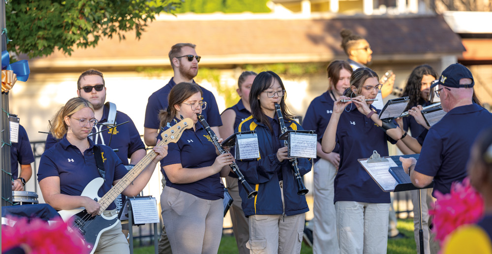 Members of the Wilkes University marching band, including a bassist, clarinetists, and flutists, perform outdoors in navy polo shirts while following a conductor.