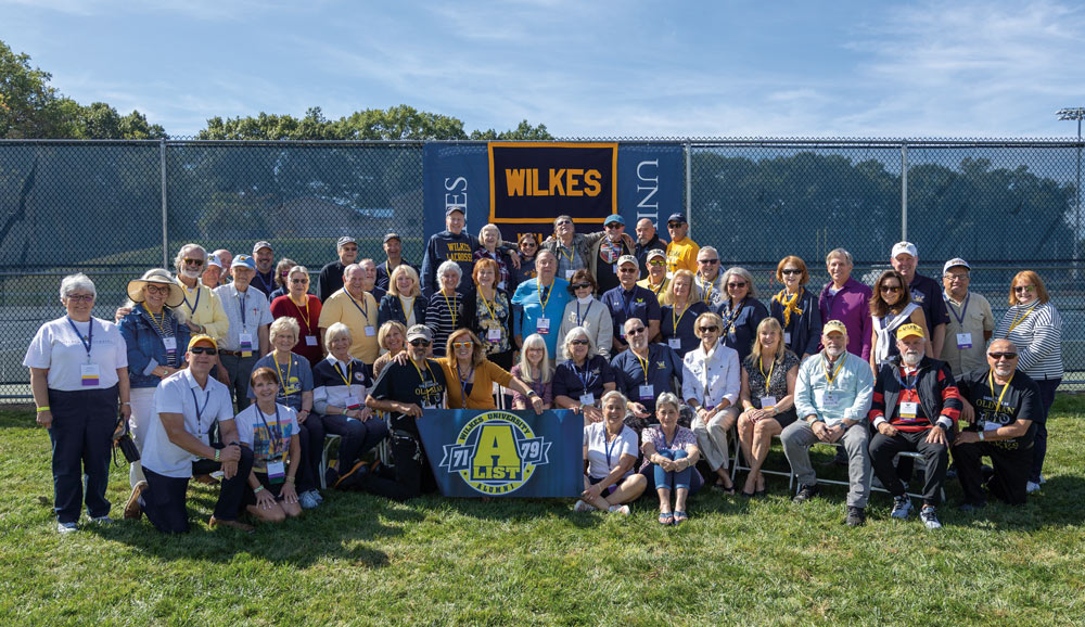 A large group of Wilkes University alumni from the classes of 1971 to 1979 pose together on a sunny field, holding a blue "A-List Alumni" banner in front of a chain-link fence.