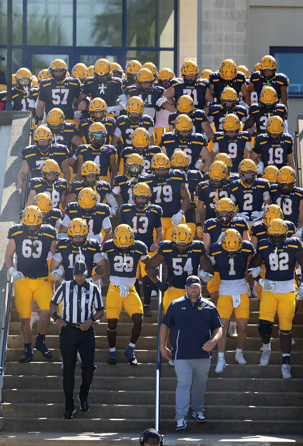 The Wilkes University football team, wearing navy jerseys and yellow helmets, walks down a concrete stadium staircase led by their coach and an official before a homecoming game.