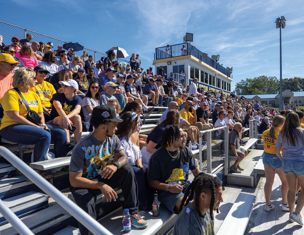 A wide shot of a crowded spectator section at the Wilkes University stadium. Fans sit in bleachers under a bright blue sky, with a white press box building visible in the background.