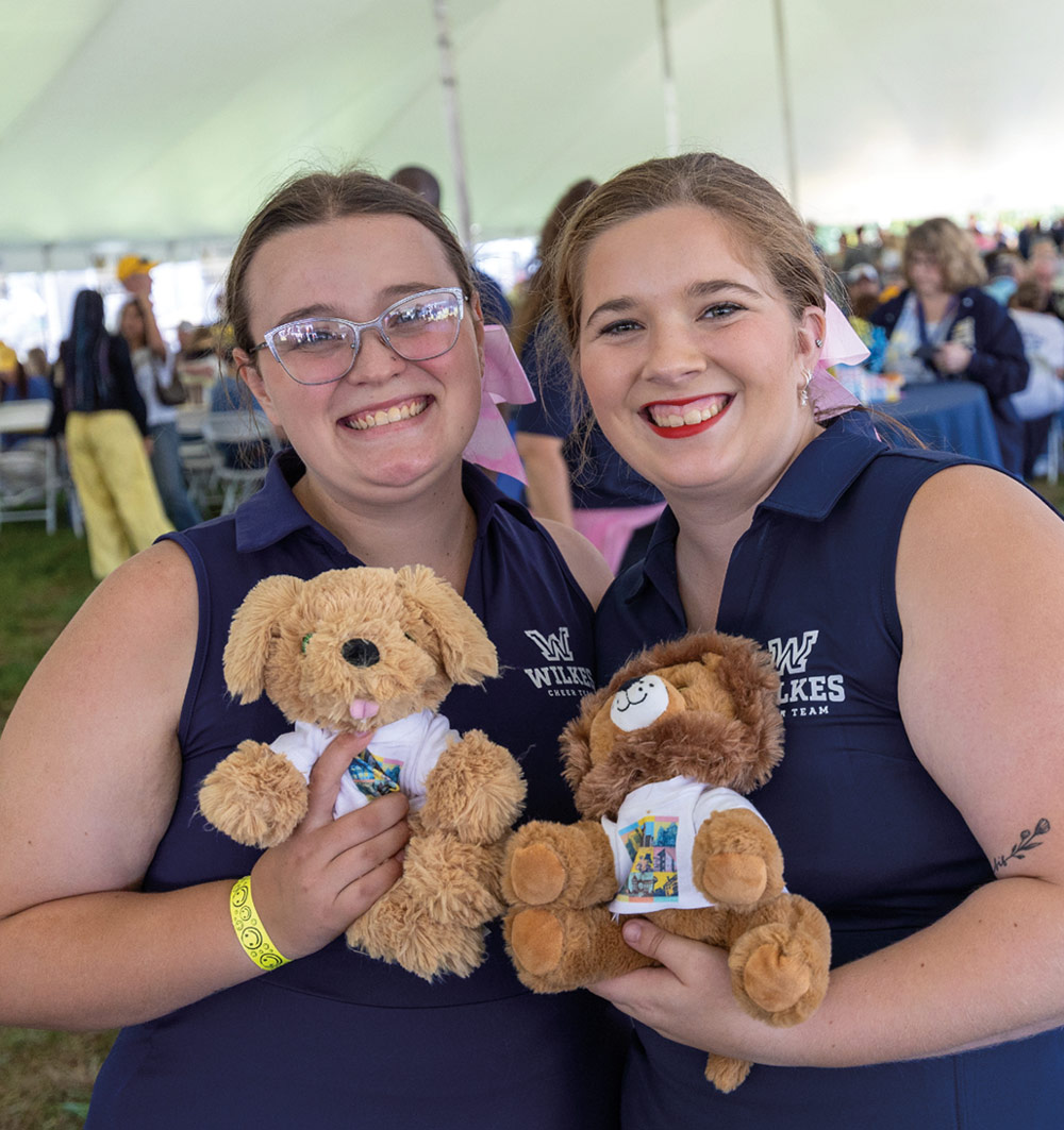 Two smiling Wilkes University students in navy blue vests pose together under a white tent, each holding a small stuffed animal wearing a tiny, colorful homecoming-themed t-shirt.