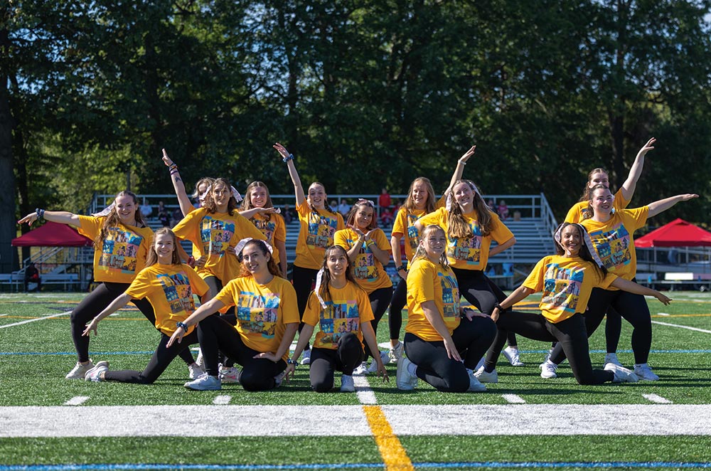 A dance team in matching gold Wilkes University t-shirts and black leggings performs a choreographed routine on the green athletic turf during a homecoming event.