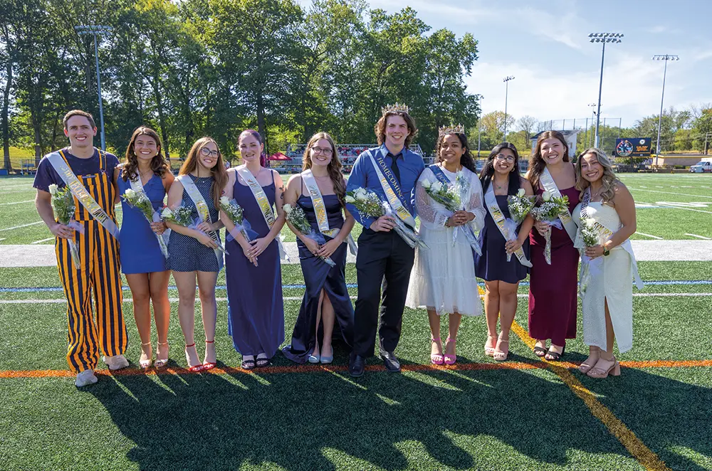 The Wilkes Homecoming Court poses together on the athletic field. The students wear formal attire and sashes, with the king and queen wearing crowns and holding bouquets.