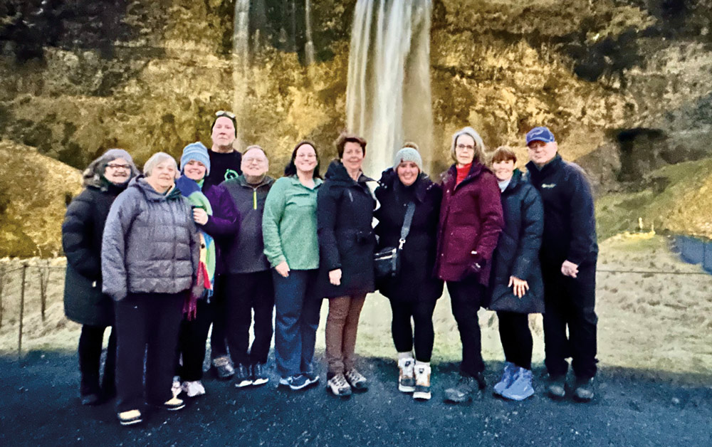 A group of eleven travelers in winter coats and hats pose for a photo in front of a tall, misty waterfall cascading down a rocky cliffside during a group excursion.
