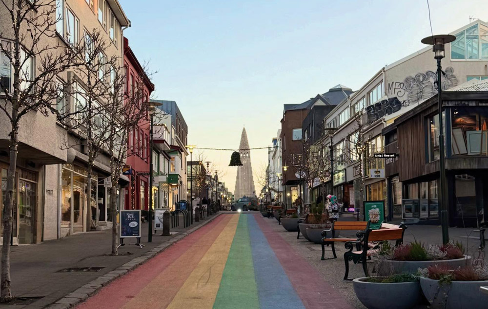 A street view of Reykjavik, Iceland, featuring a rainbow-painted road leading toward the Hallgrimskirkja church, with shops and cafe seating lining both sides of the path.