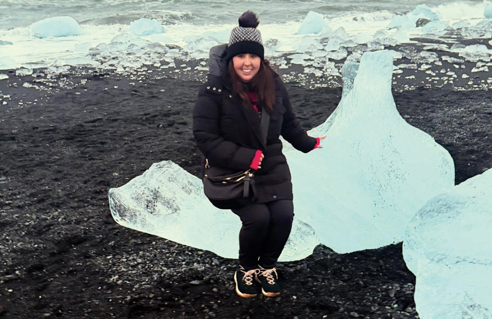 A woman in a black puffer coat and winter hat poses on a large piece of crystal-clear glacial ice on a black sand beach, with smaller ice fragments and ocean waves behind her.