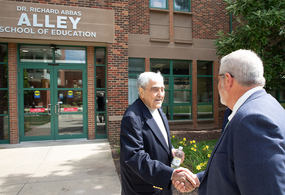 Dr. Alley shaking hands with a man, in front of the Dr. Richard Abbas Alley School of Education building sign.