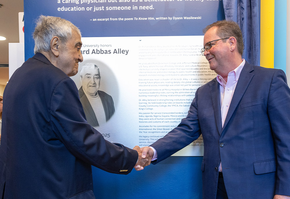 Dr. Alley shaking hands with another man in a blue suit, in front of a blue wall with biographical information and a photo.