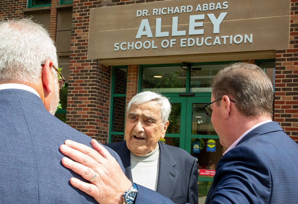A group of people from behind, looking at Dr. Alley. One man has his hand on the back of another man. The building sign is in the background.