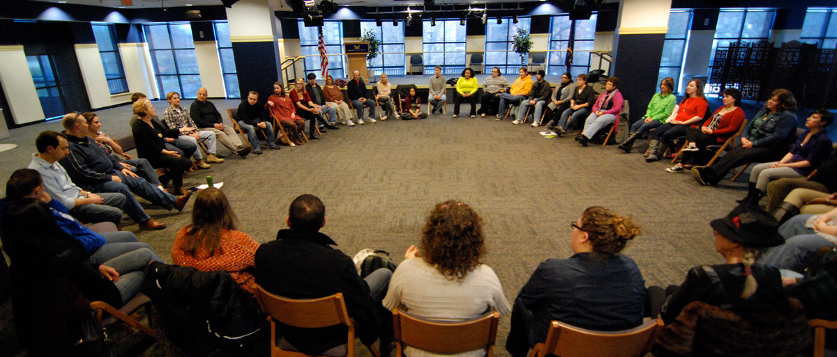 A large group of people sit in a wide circle of chairs for a discussion in a spacious room with floor-to-ceiling windows.