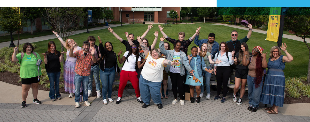 A diverse group of students and faculty pose with arms raised on a paved campus walkway. In the background is a large green lawn and the Lawrence & Sally Cohen Science Center building.