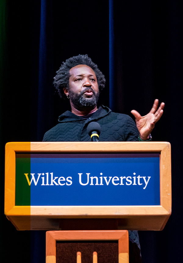 Marlon James speaks in to a microphone as he stands behind a podium bearing the Wilkes University name