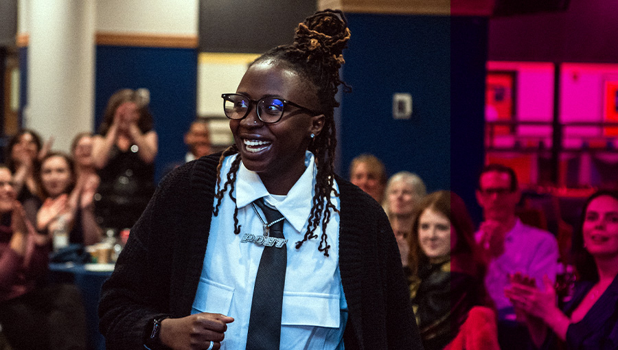 an individual with loc'd hair smiles as the walk down an aisle between audience filled seats